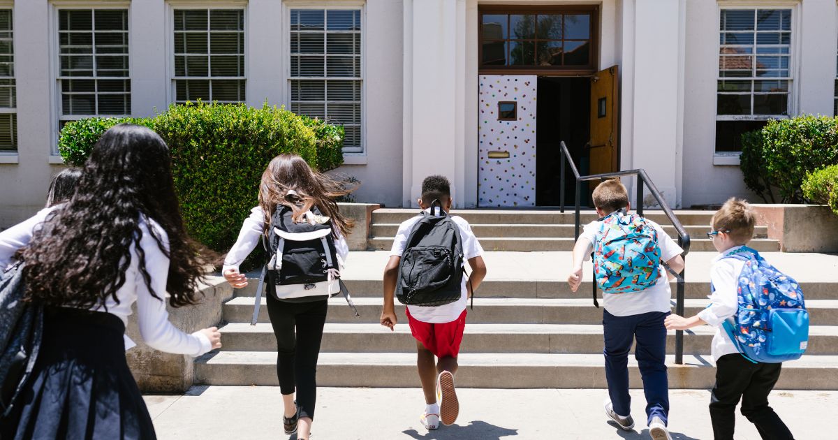 Five children with backpacks running up the steps toward a school entrance.